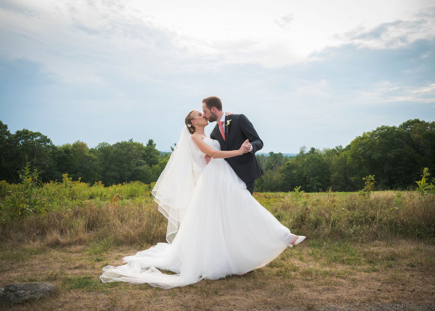 A romantic wedding couple shares an intimate dance in a scenic meadow with flowing white dress and blue sky background.