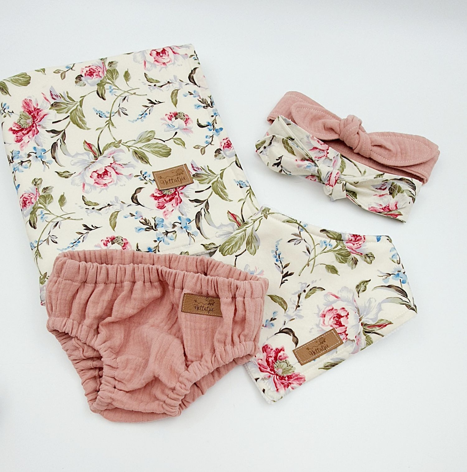 Baby girl gift set with floral swaddle blanket, pink bloomers, headband bow, and bandana bib on white background.