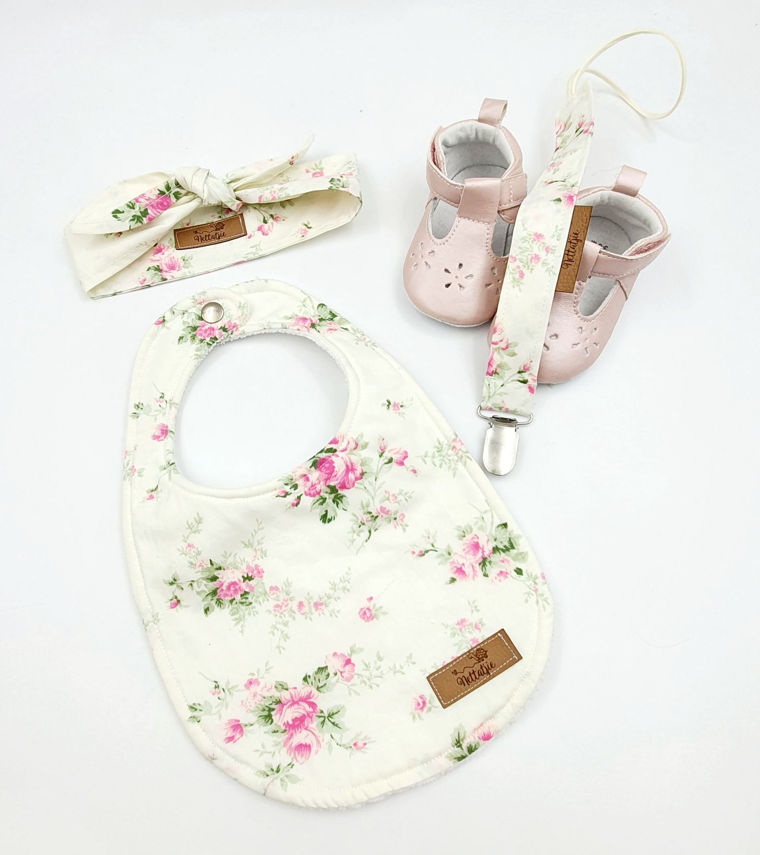 Baby girl gift set with floral bib, headband, pink Mary Jane shoes, and pacifier clip on white background.