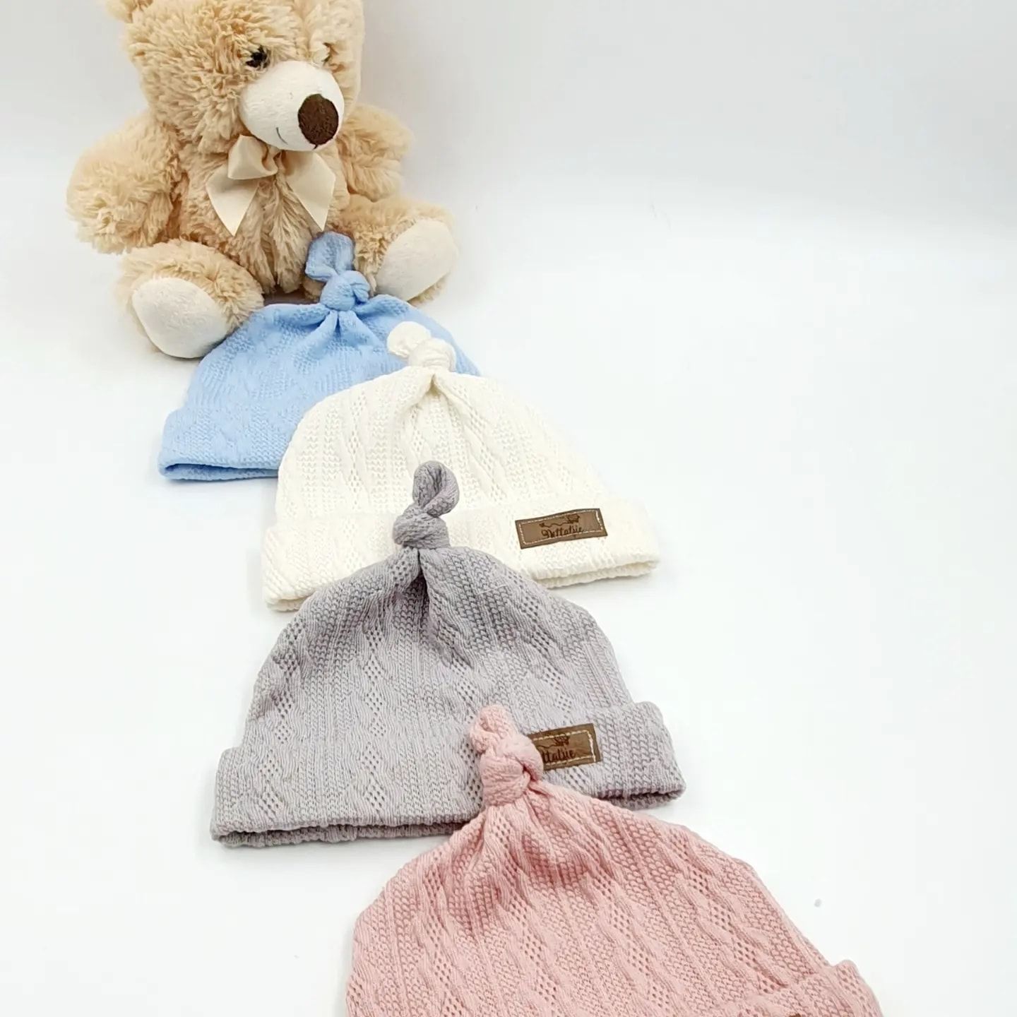 Four knot-top baby beanies in blue, white, gray, and pink displayed with a teddy bear.