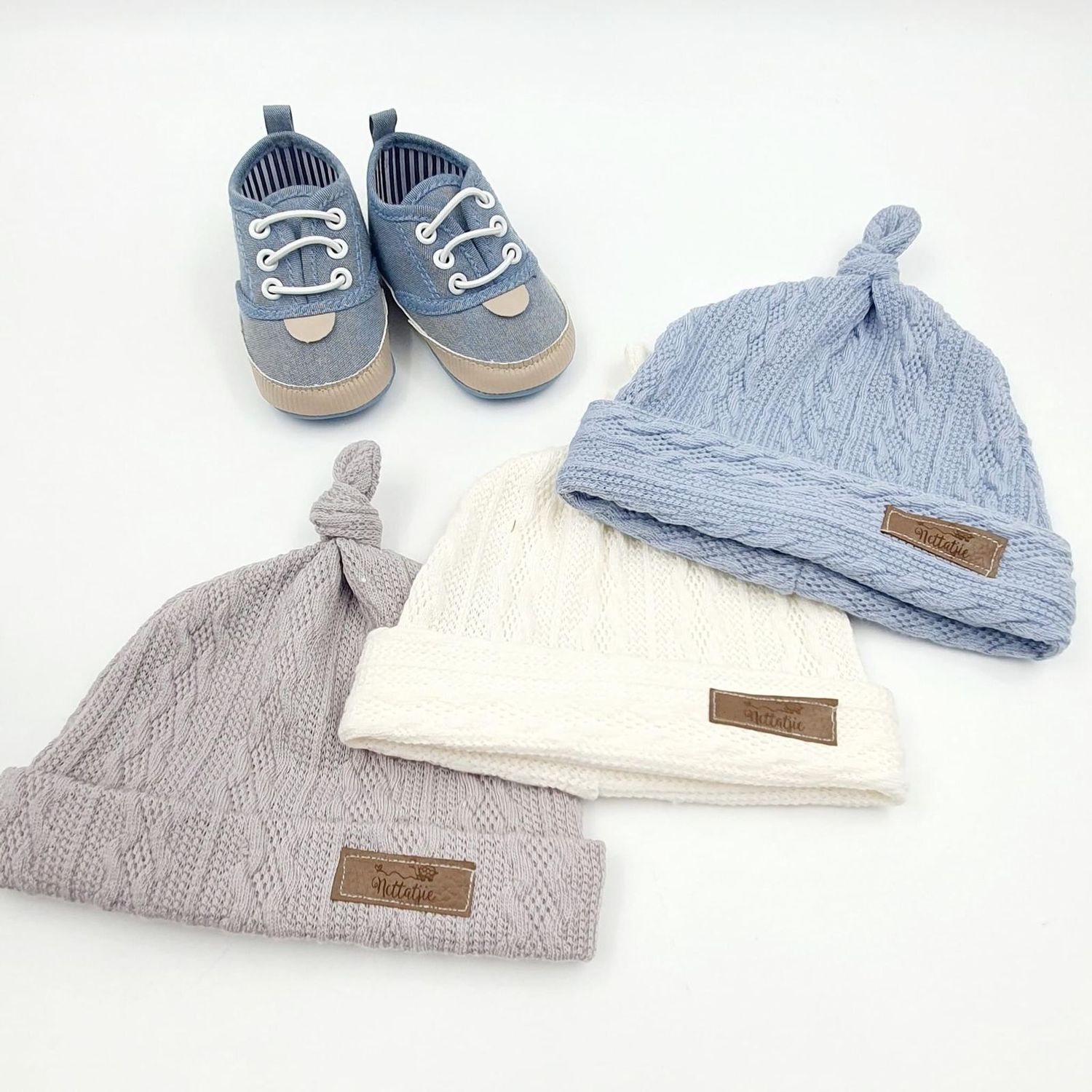 Baby blue sneakers with laces alongside three knit beanies in blue, white, and gray with knotted tops on white background.