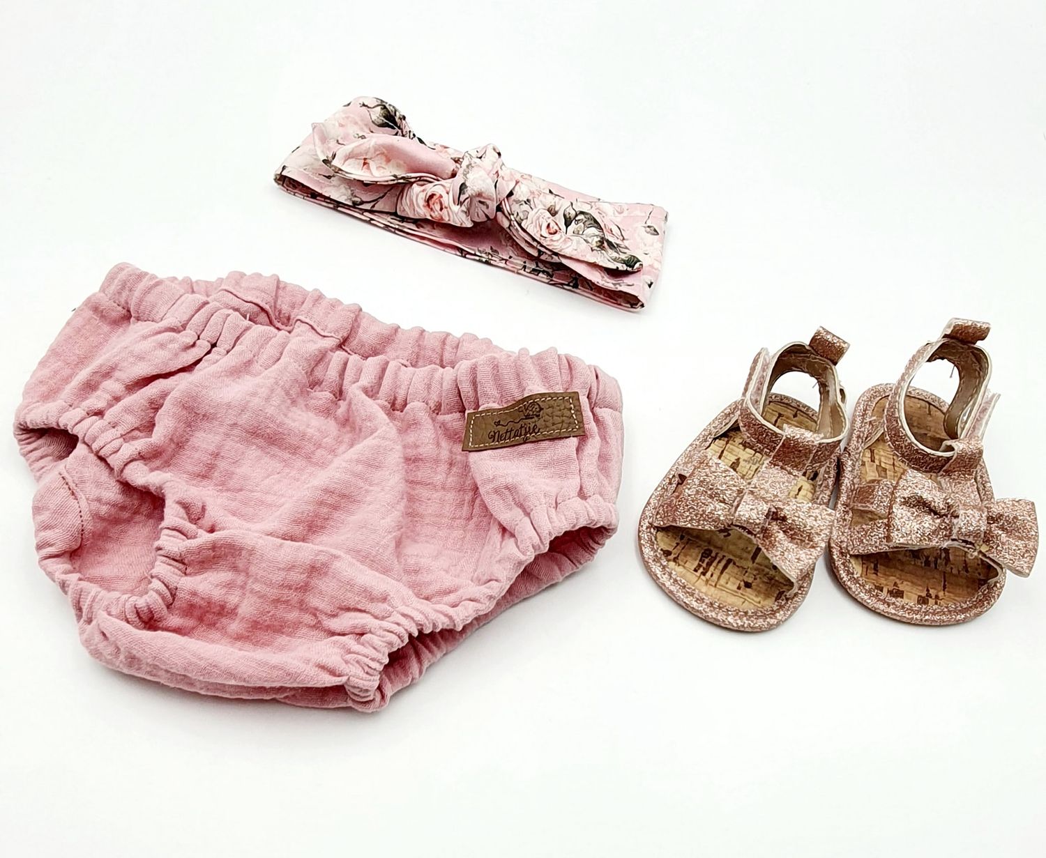 Pink muslin baby bloomers set with floral knotted headband and matching cork bow sandals on white background.