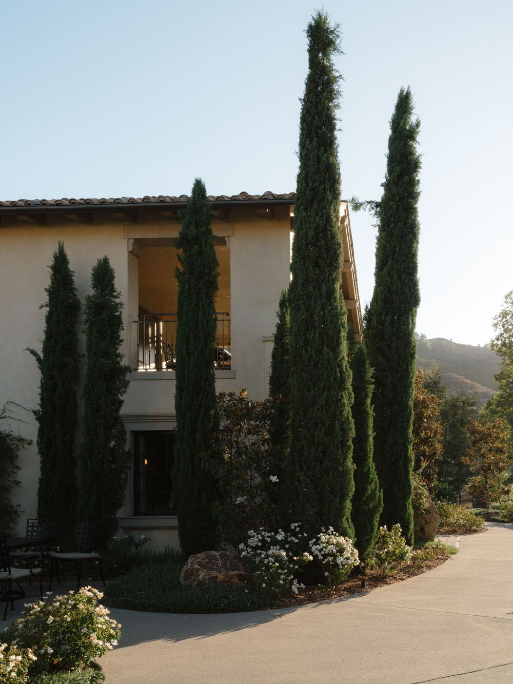 Tall Italian cypress trees line a Mediterranean-style villa with stucco walls, balcony, and mountain views in the background.