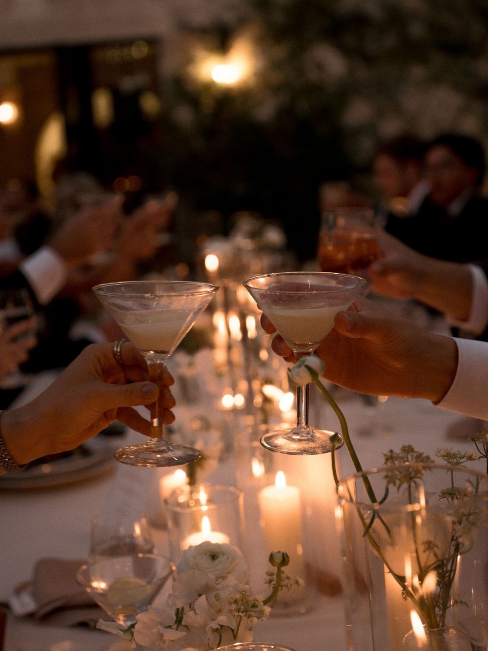 Guests toasting with martini glasses at a candlelit wedding reception dinner table adorned with white flowers.