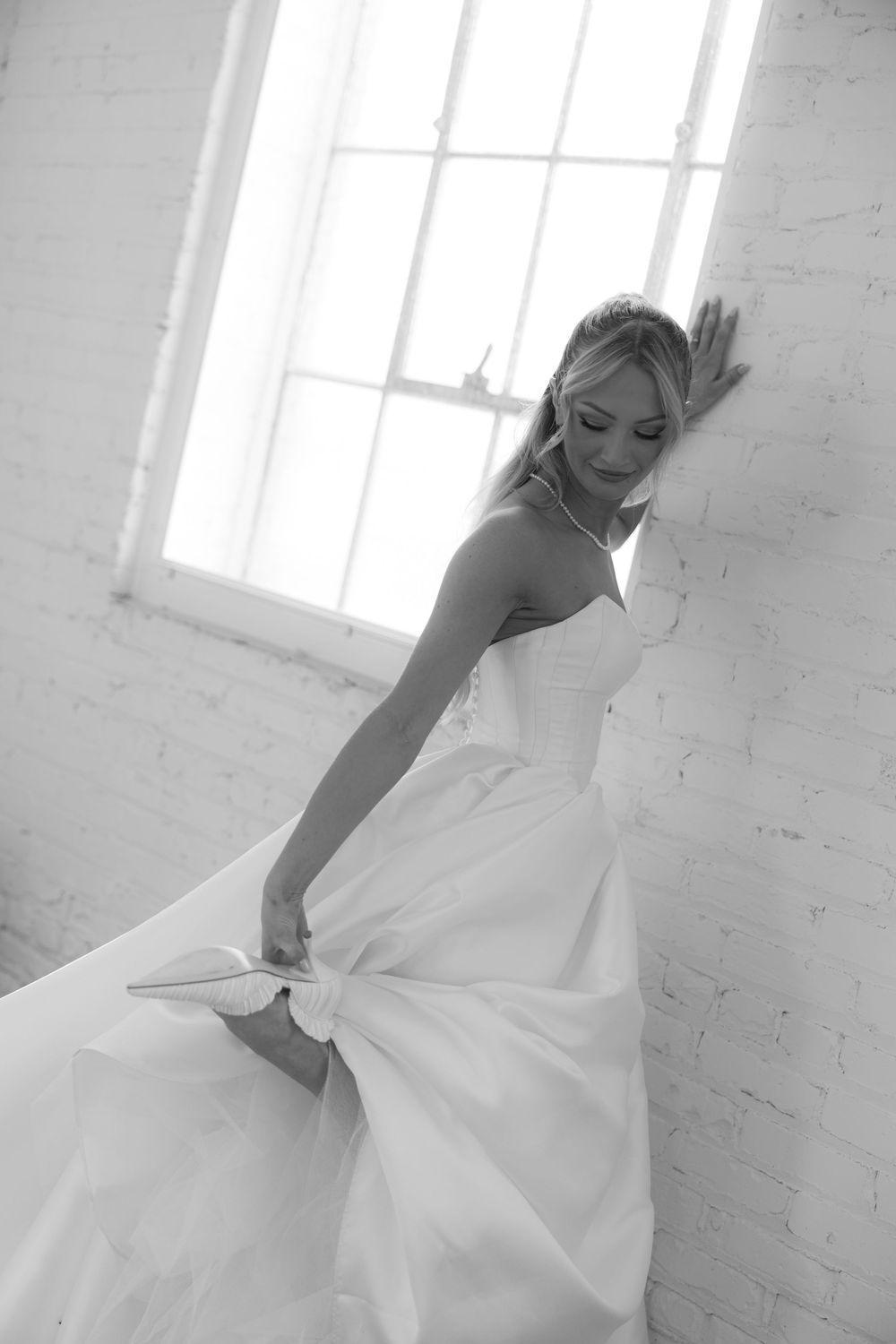 Romantic black and white photograph showing a bride in a flowing white gown twirling beside a bright window.