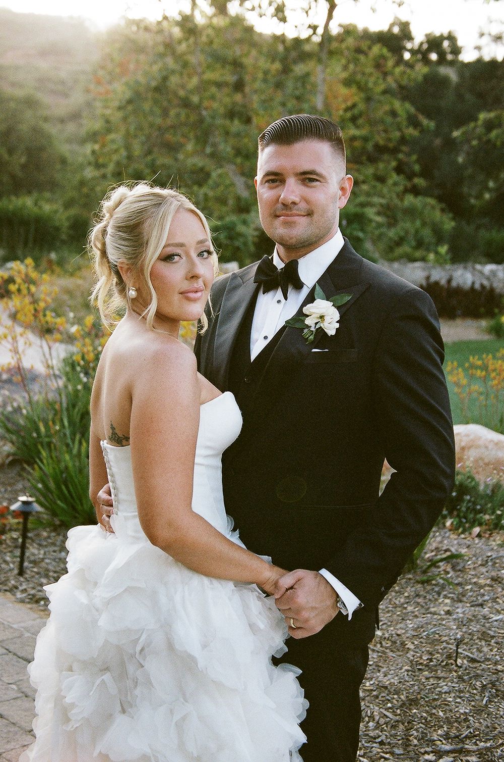 A bride in a white layered gown and groom in black tuxedo pose together at sunset in an outdoor garden setting.