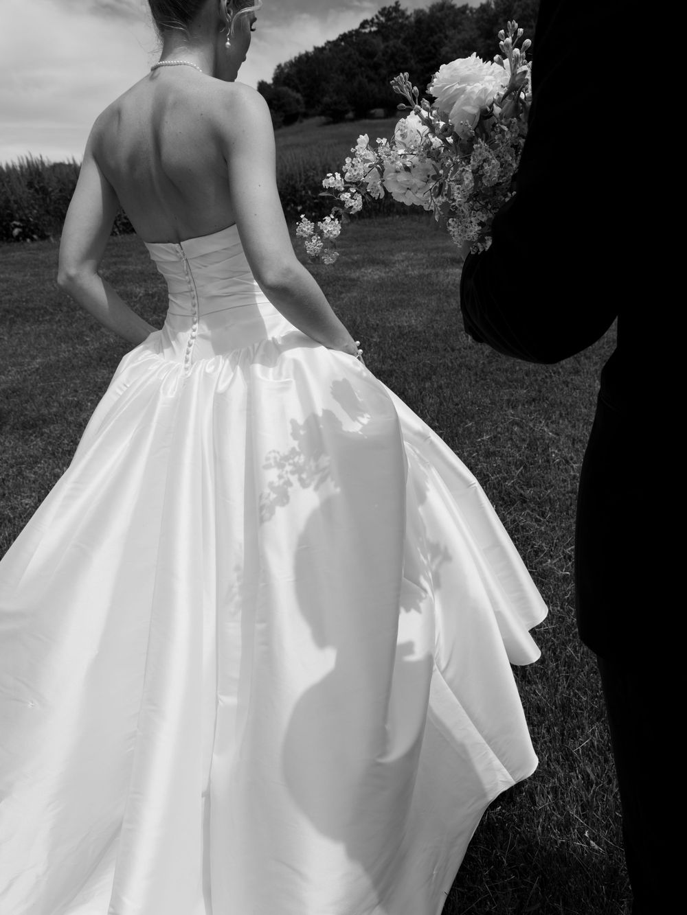 Black and white photo of bride in strapless ball gown holding bouquet with groom silhouette outdoors.
