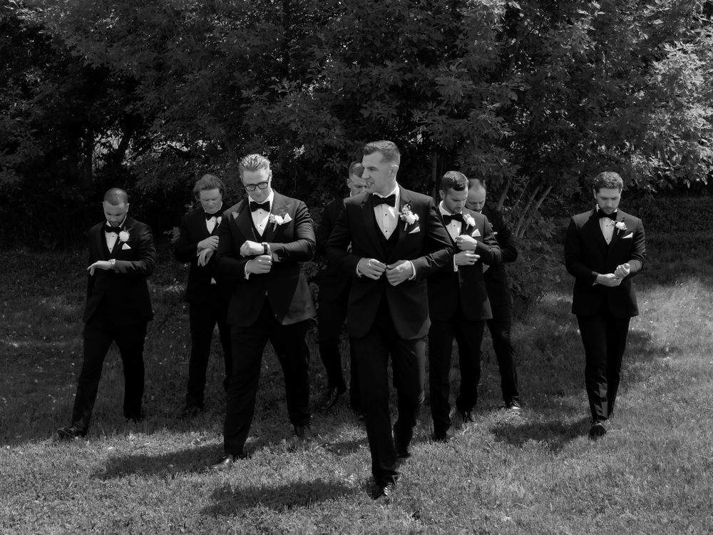 Groom and groomsmen in black tuxedos walking confidently outdoors in a black and white wedding photo.
