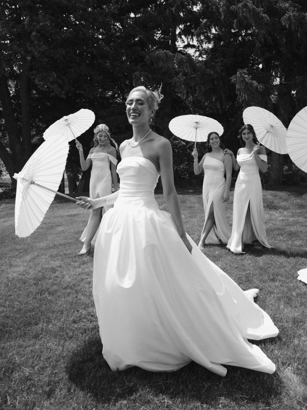 Bride in white gown twirls outdoors while bridesmaids hold parasols in black and white wedding photo.
