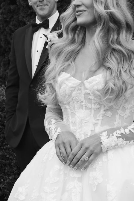 Black and white photo of bride in lace gown with engagement ring and groom in tuxedo at wedding ceremony.