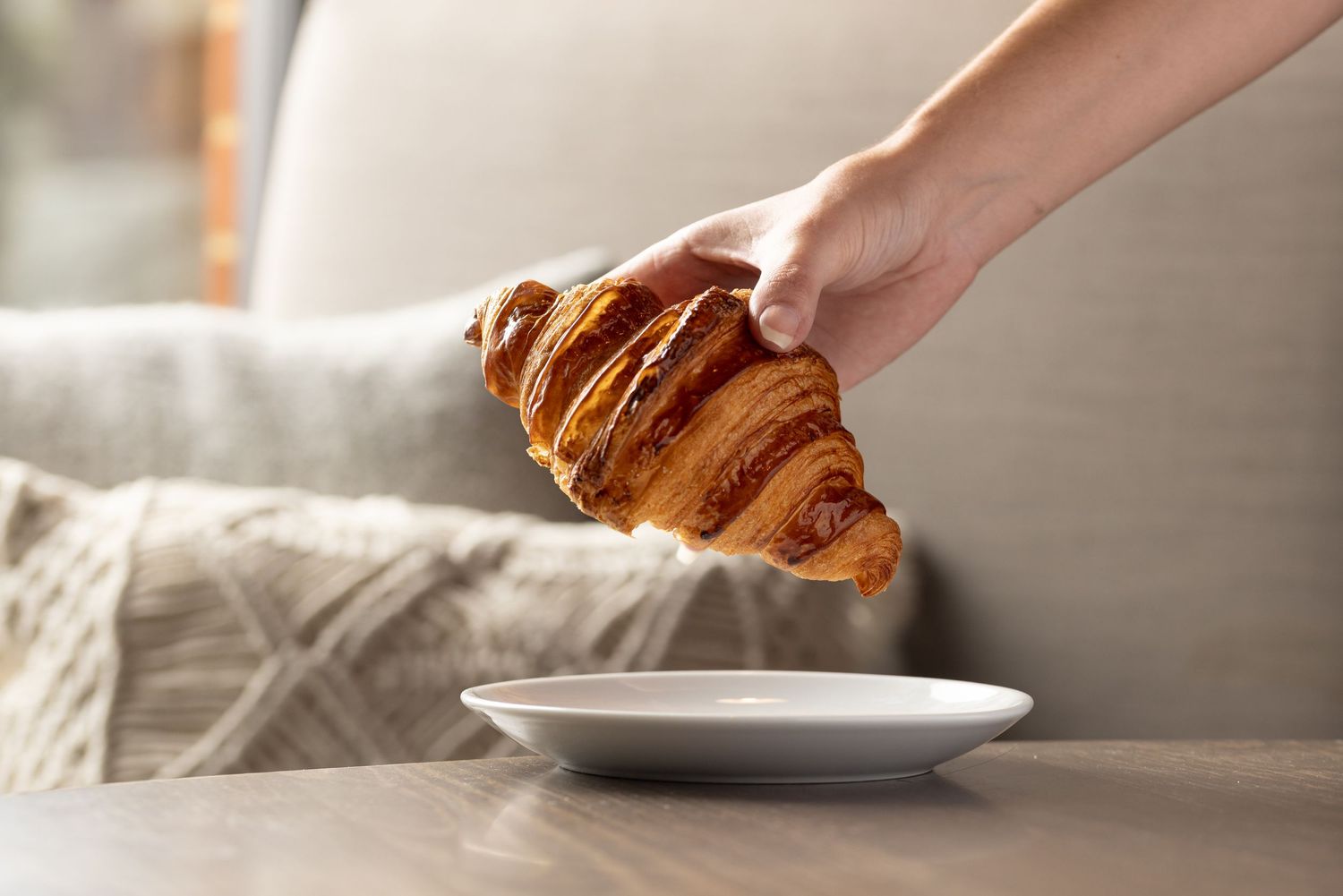 Hand placing a golden flaky French croissant onto a white plate in natural light, bakery cafe brunch food photography Southampton