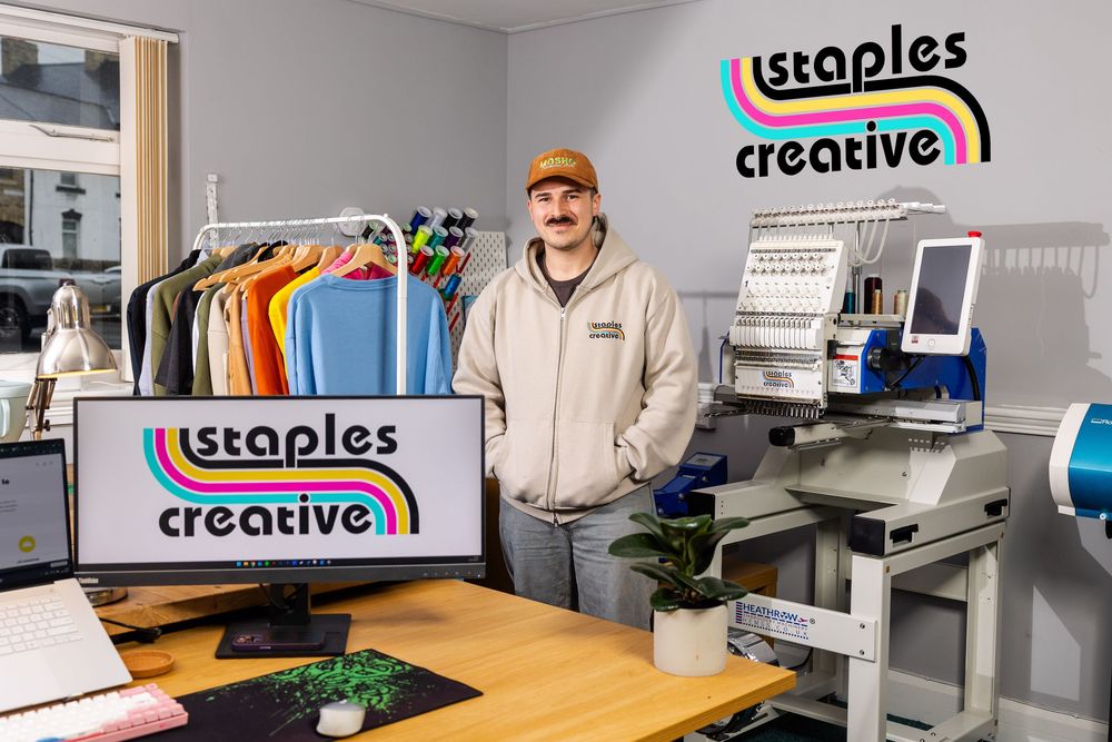 Man standing in a colourful embroidery studio with branded “Staples Creative” signage, clothing rack, and equipment in the background.