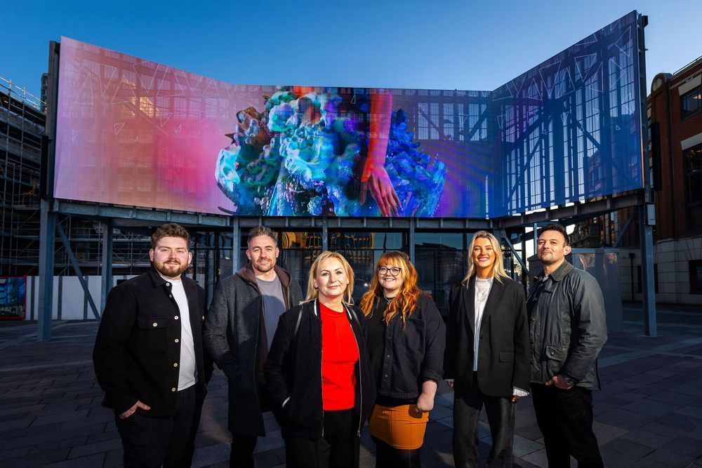 Group portrait of six people standing outdoors in front of a large curved digital screen displaying vibrant artwork.