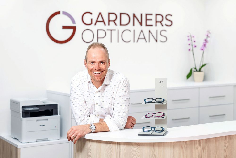 Professional business portrait of an optician at reception with eyewear display in a bright, modern optical practice.