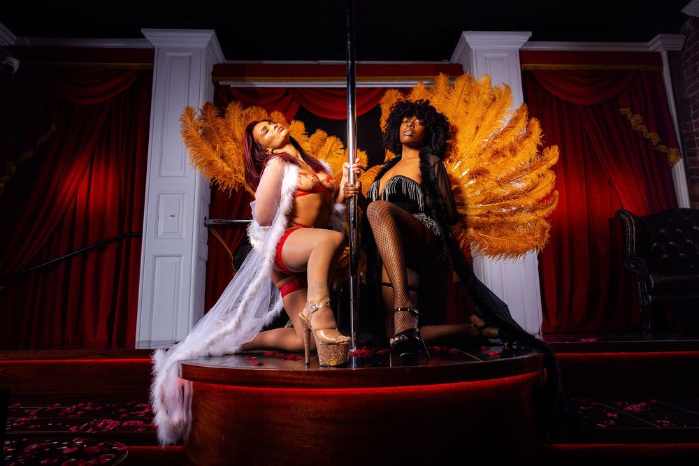 Two performers posing on a stage with feathered wings and dramatic lighting in a cabaret-style setting.