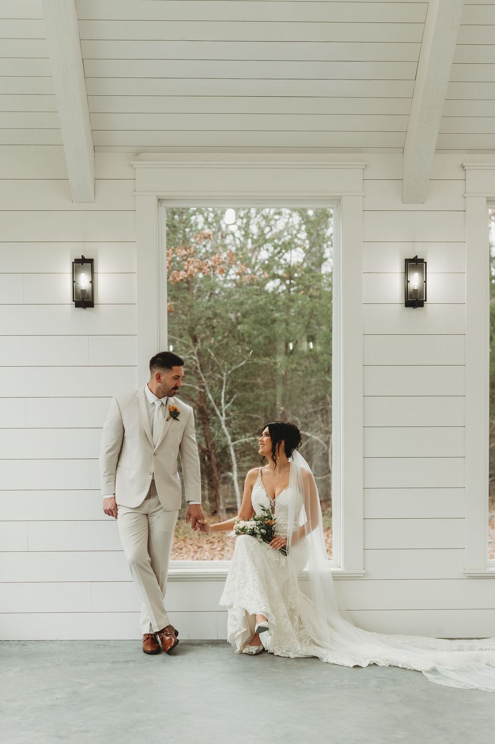 A romantic wedding portrait of a couple on a white porch with lantern sconces and a natural backdrop.