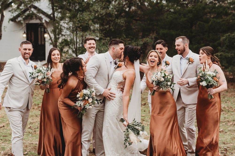 Wedding party members in brown and beige attire laugh together while holding floral bouquets outdoors.