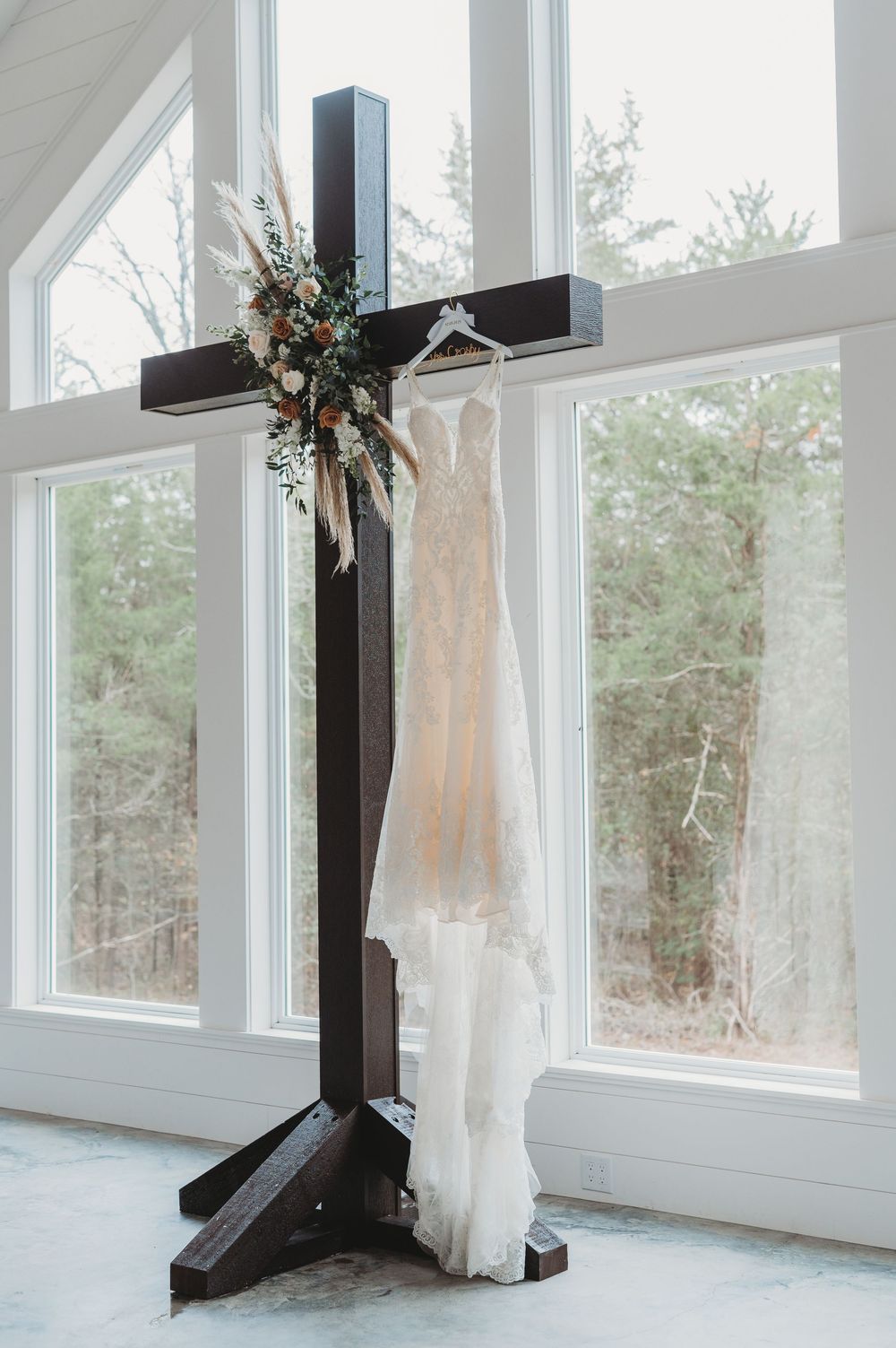A white wedding dress hangs elegantly on a dark wooden cross stand by large windows overlooking a forest view.