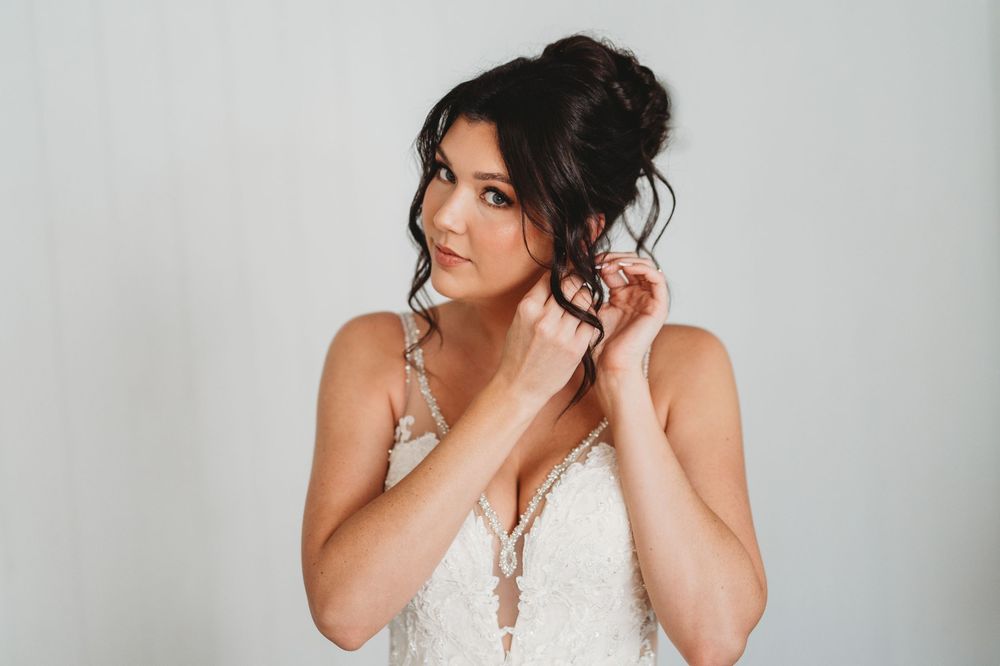A bride in a lace wedding dress adjusts her hair while posing against a light gray background.