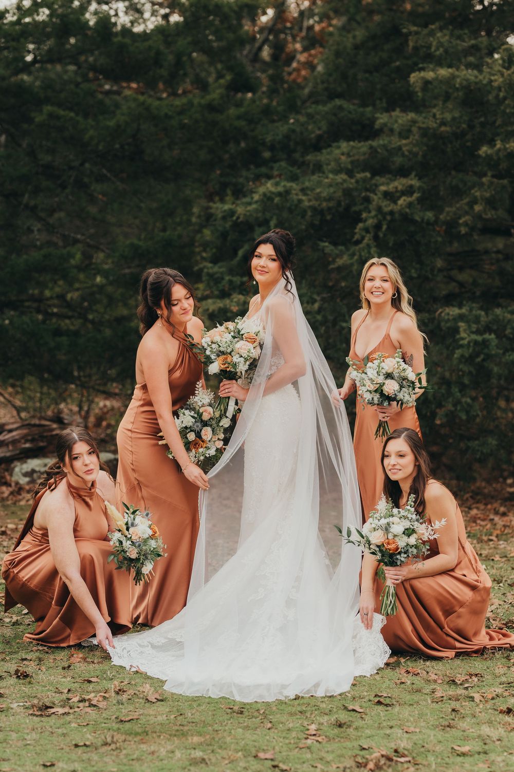 Bride in white gown sits on grass surrounded by bridesmaids wearing rust-colored dresses holding floral bouquets.