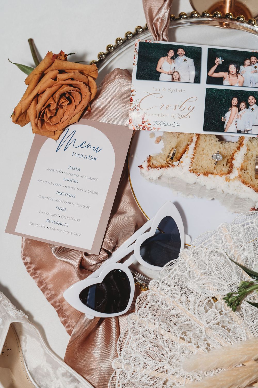 Wedding details flatlay with lace, invitations, dried flowers and photos in soft blush and neutral tones.