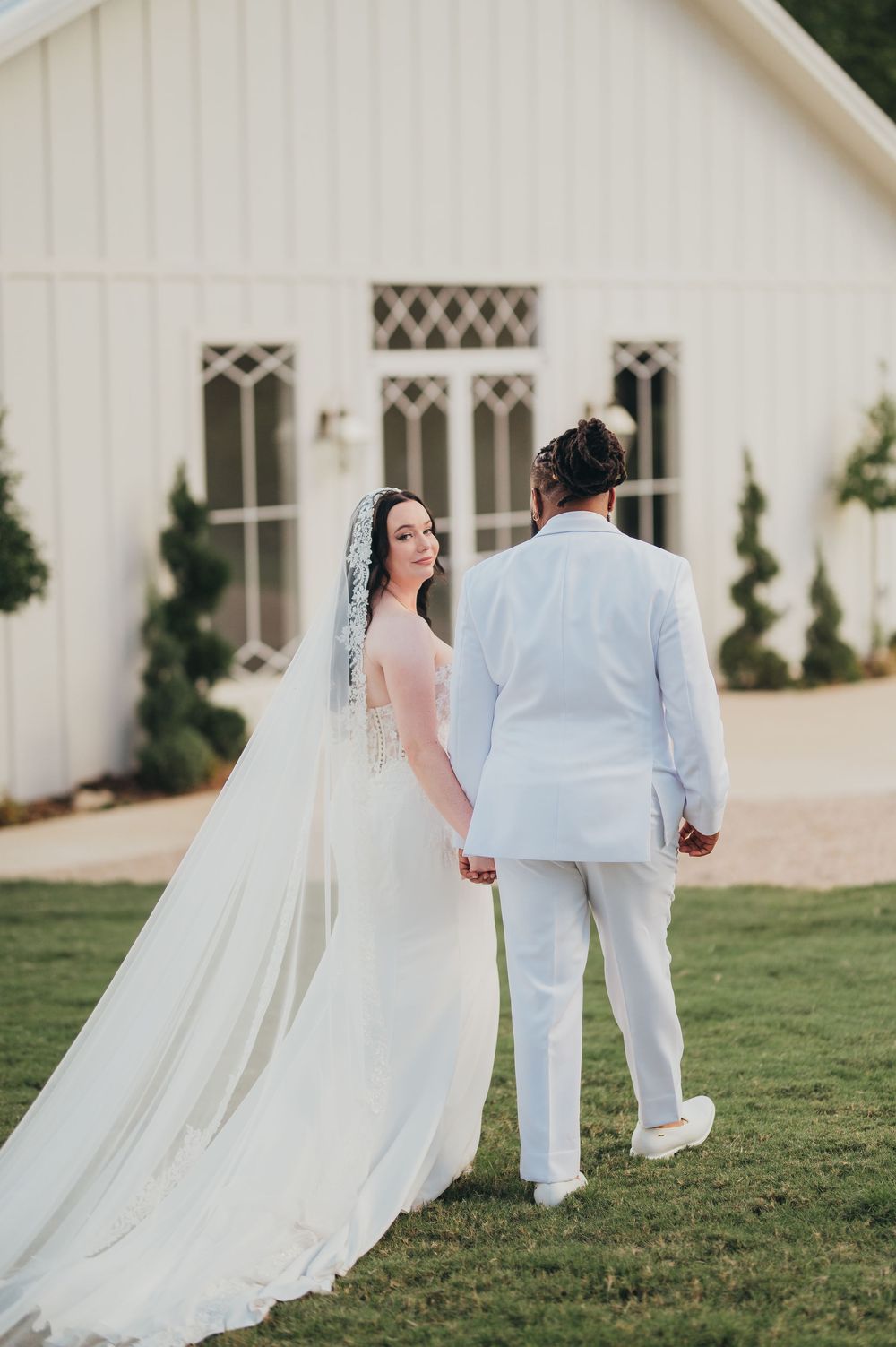 A bride in a flowing white dress and veil stands with a groom in white suit outside a chapel with gothic windows.