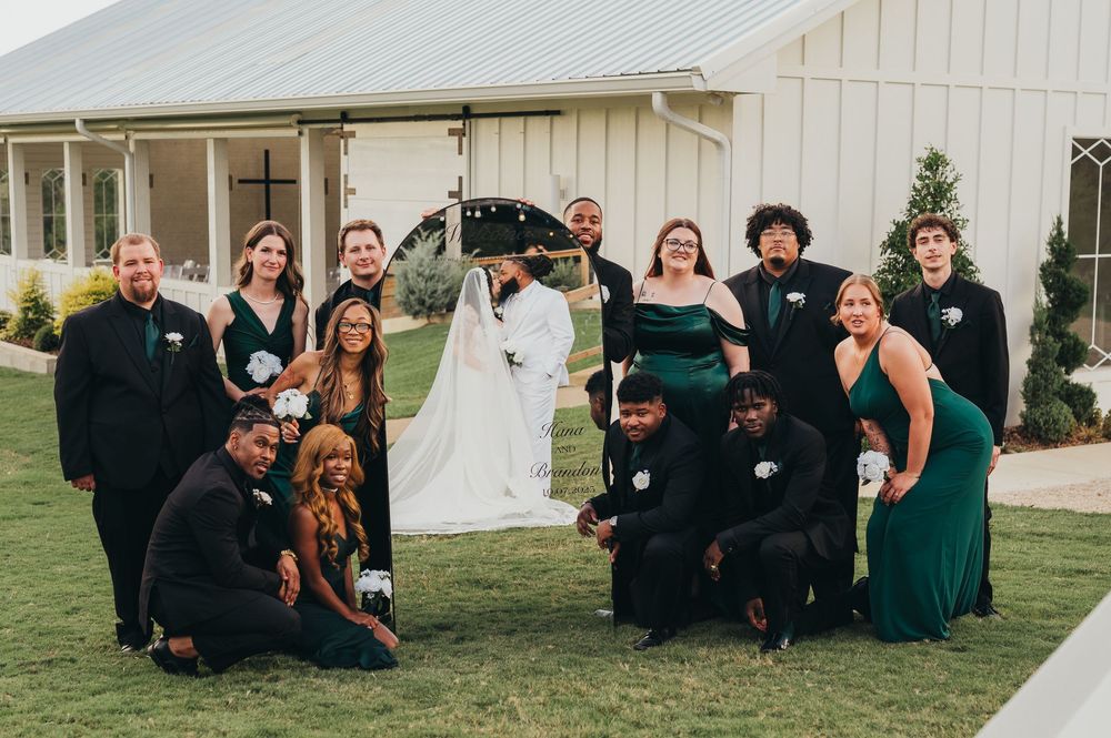 Wedding party members in black and dark green attire pose together on grass near white tent and building.