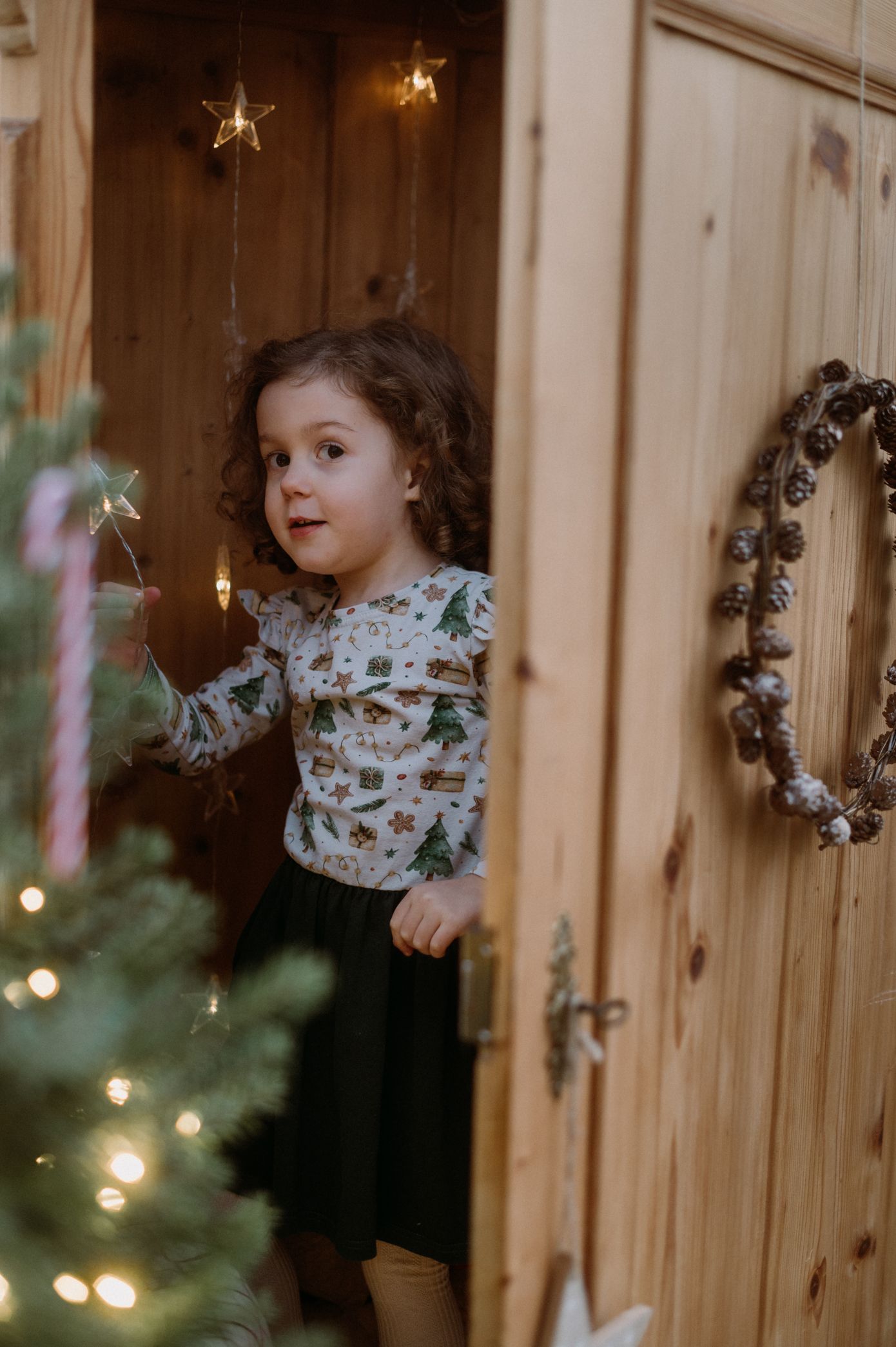 Child stands by wooden door wearing floral top and dark skirt with Christmas lights and decorations visible in background.