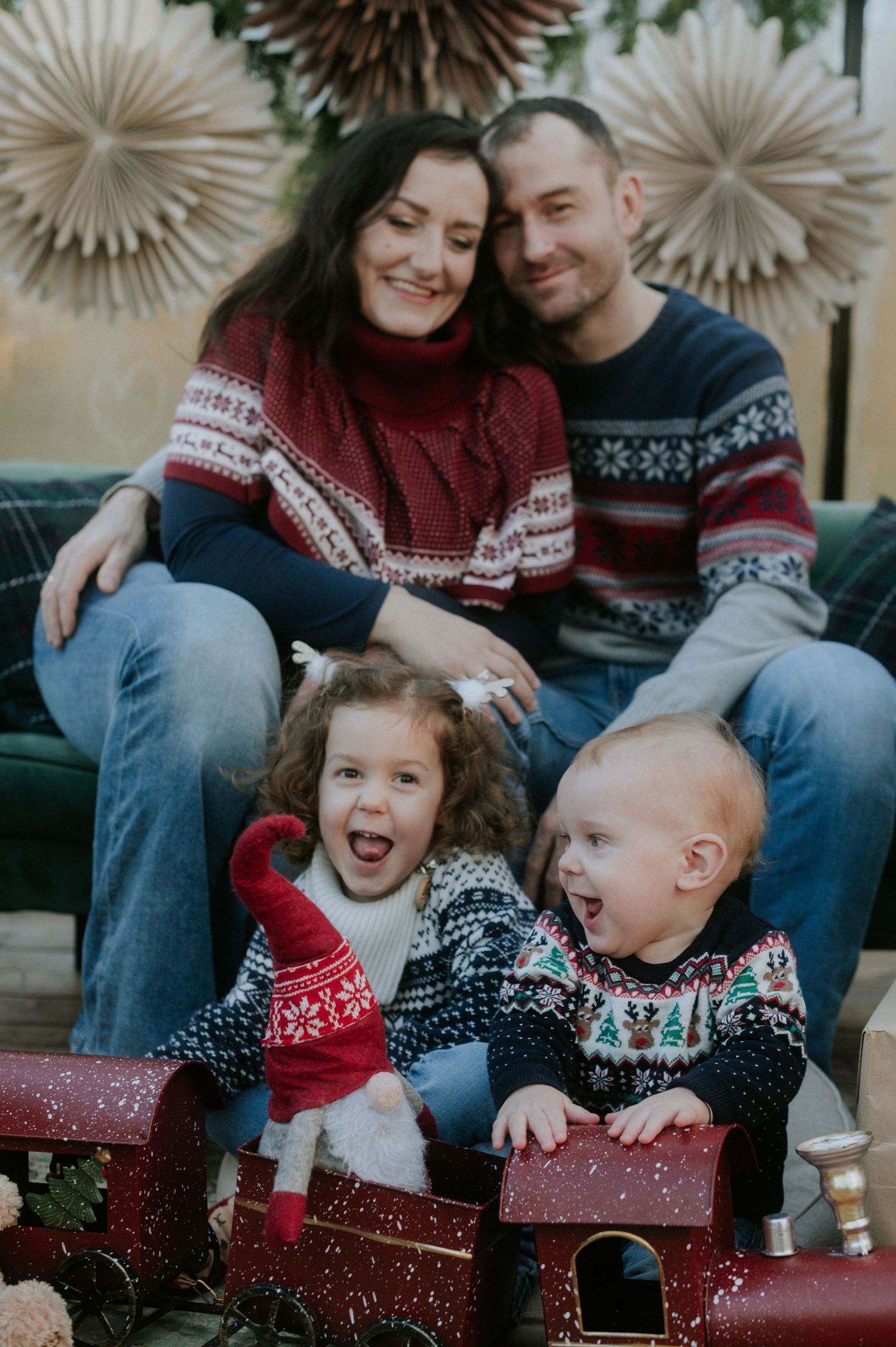 Happy family in matching holiday sweaters laughs together while opening Christmas presents on couch.