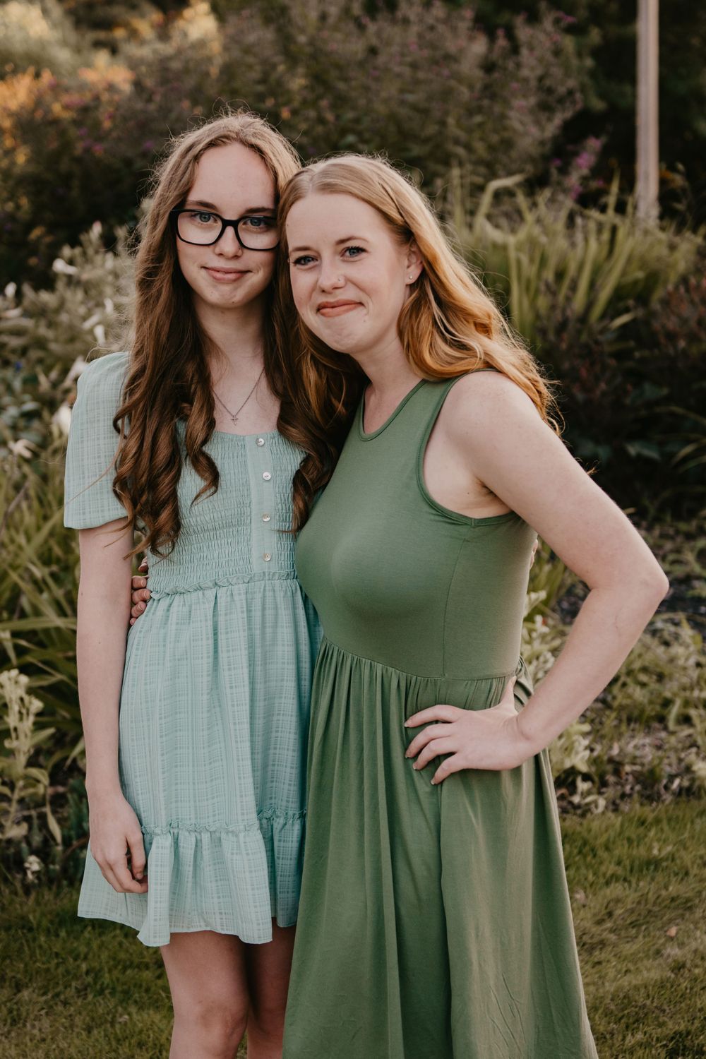 Two young women with red hair pose together outdoors in coordinating green dresses in a garden.