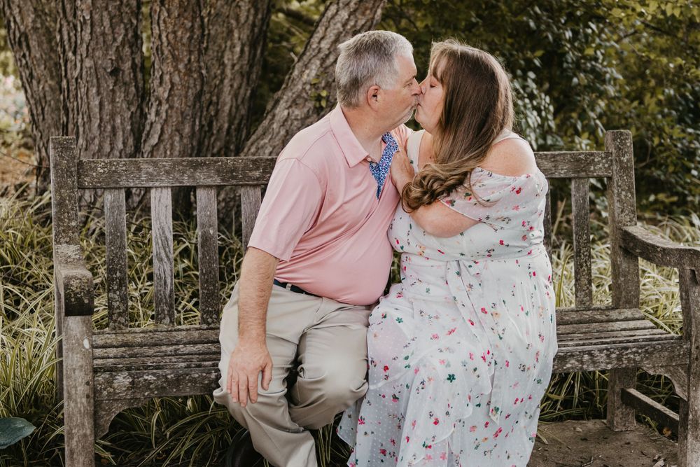 Couple shares a kiss while seated on a rustic wooden garden bench beneath a large tree.