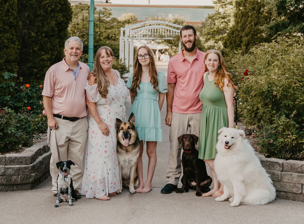 Family of six poses outdoors with four dogs including a German Shepherd and white Samoyed in a garden setting.