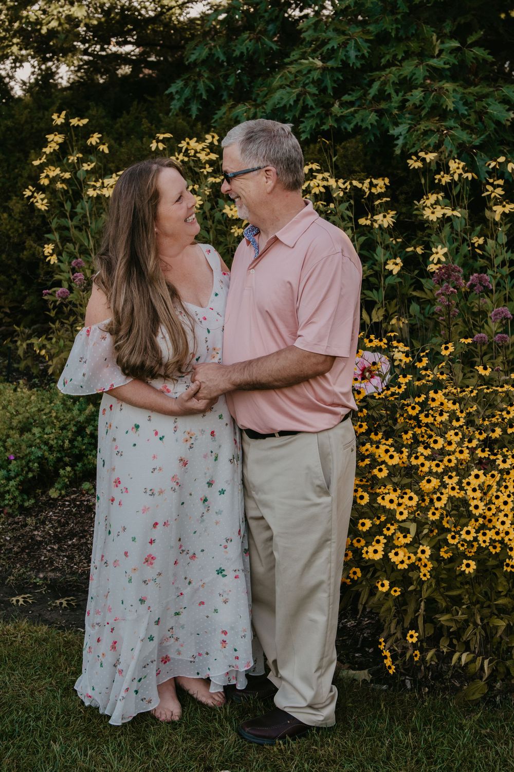 Smiling couple embraces in front of vibrant yellow black-eyed Susan flowers in a lush garden setting.