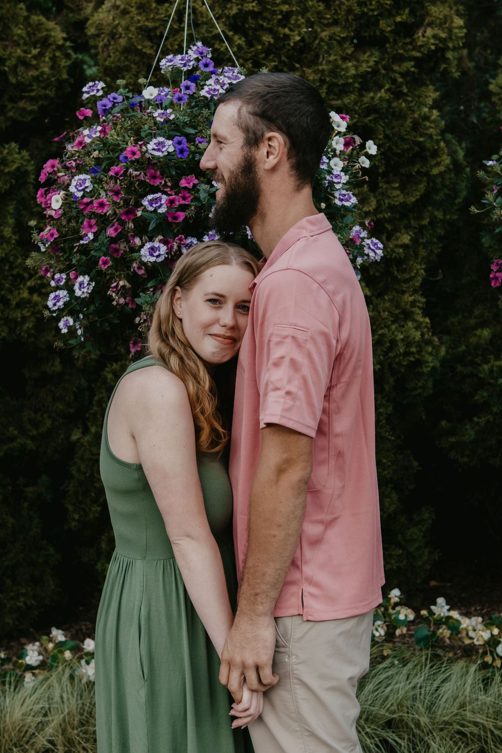Couple embraces tenderly in front of vibrant hanging flower basket with purple and pink blooms.