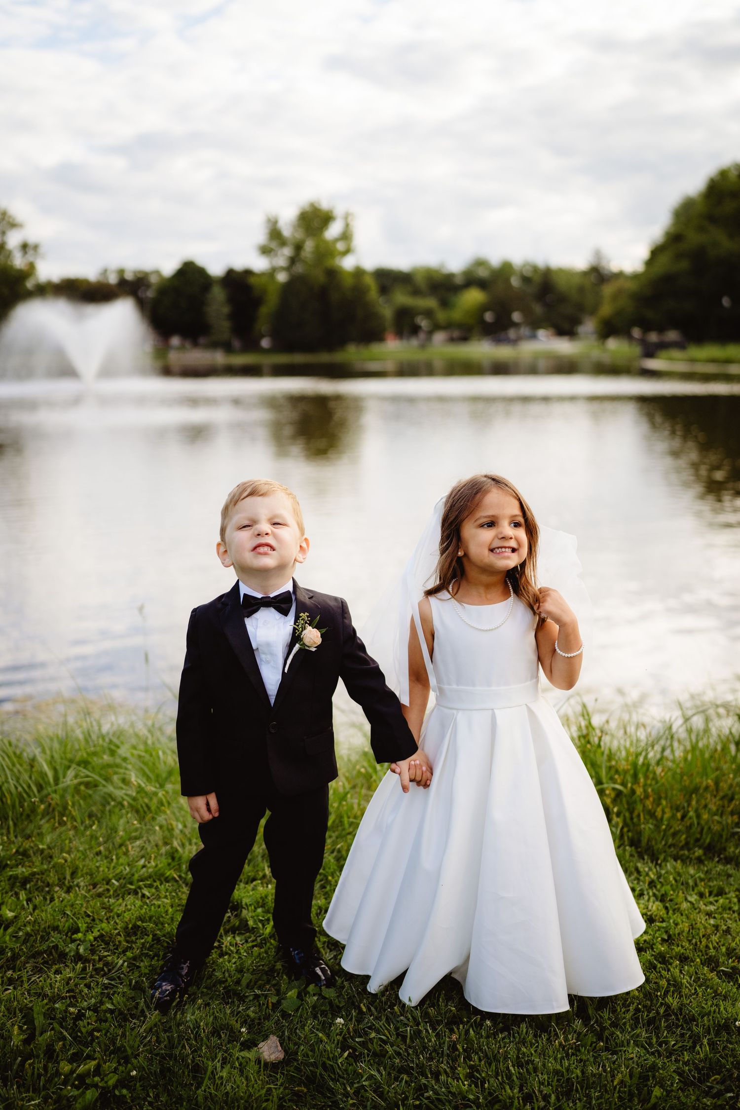 Two children in formal wedding attire stand by a lake with a fountain, holding hands against a scenic backdrop.
