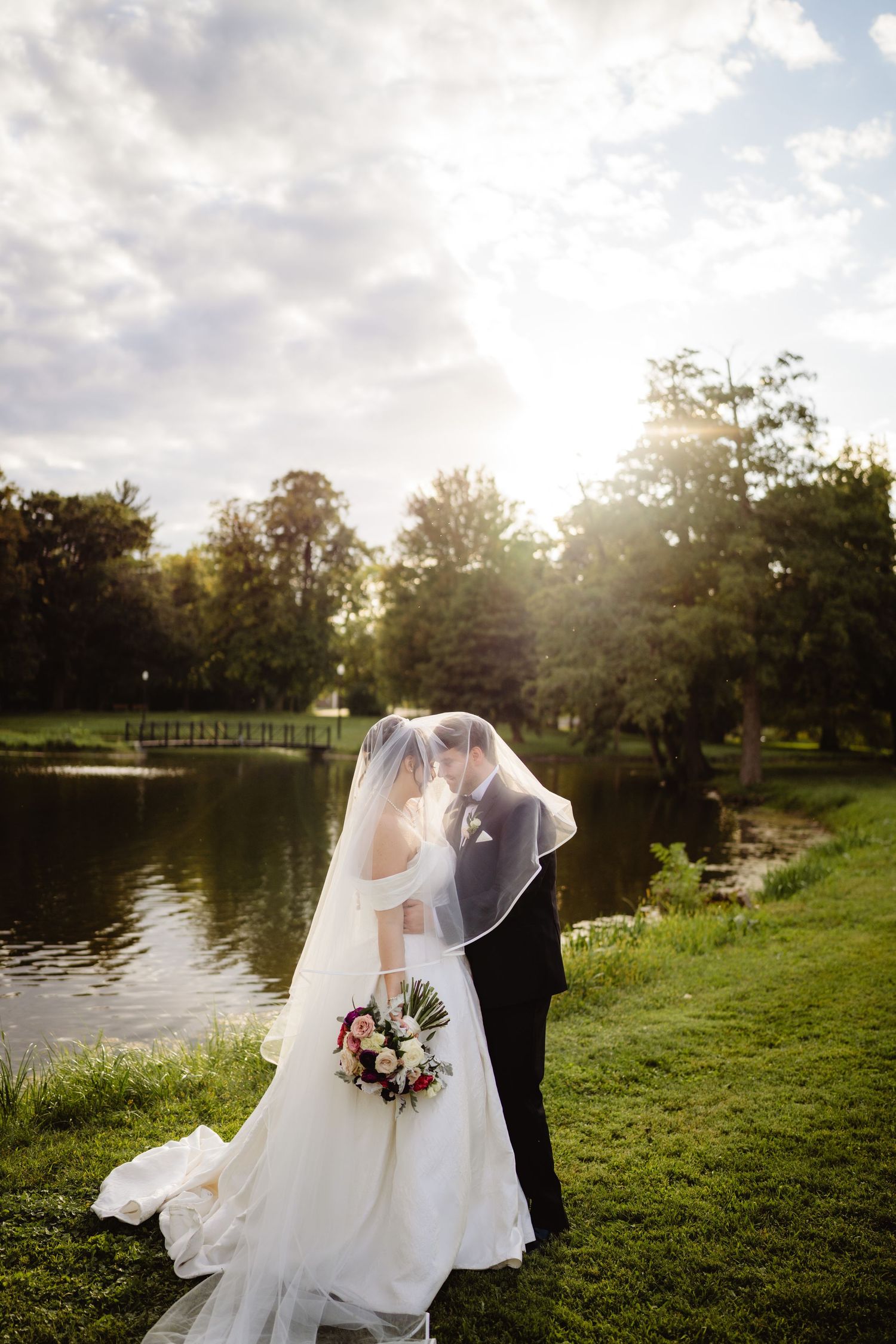 A couple in wedding attire embrace by a serene pond surrounded by trees during golden hour with sunlight peeking through.