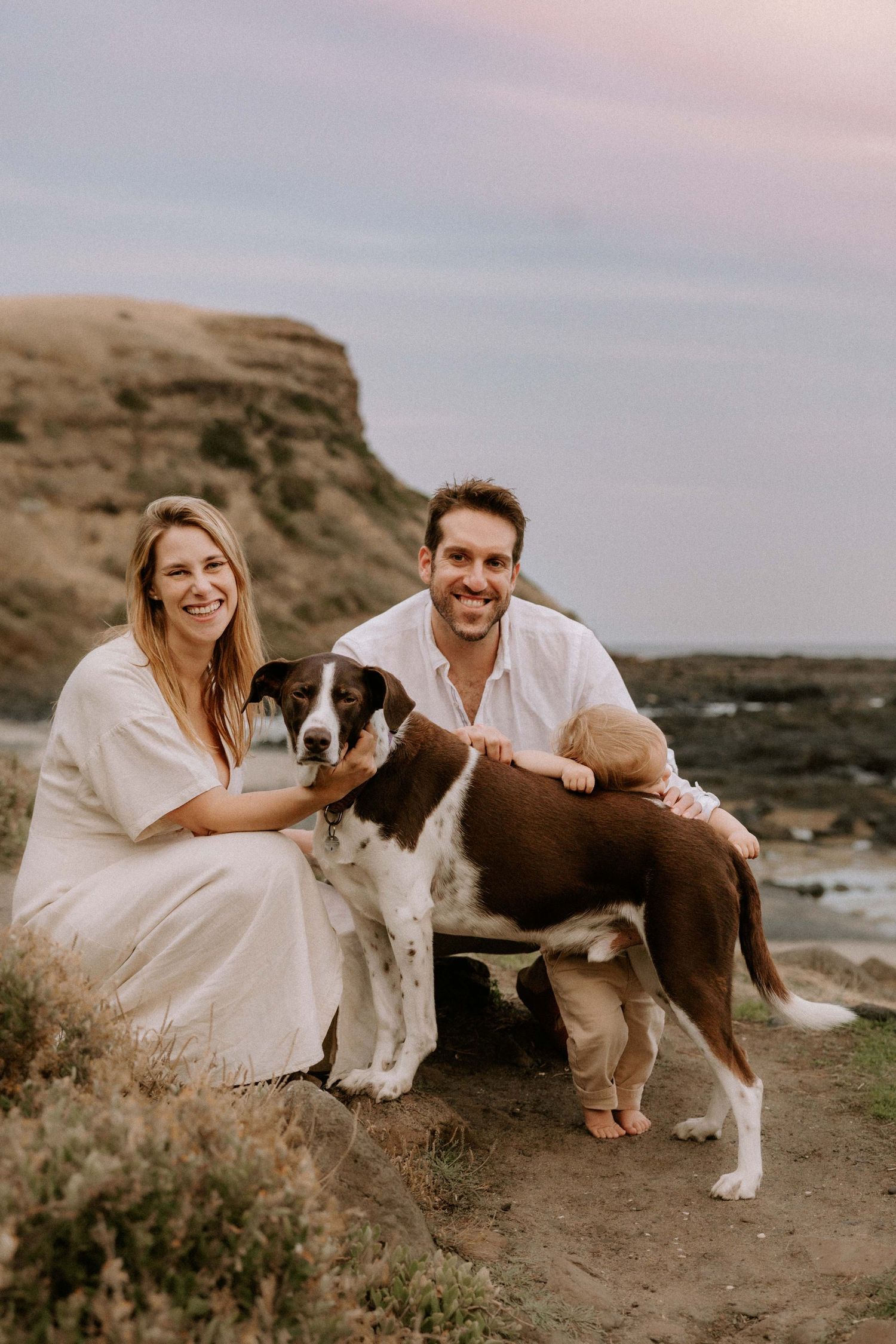 A couple in white outfits sits with their Saint Bernard dog on a rocky beach at sunset, creating a serene coastal portrait.