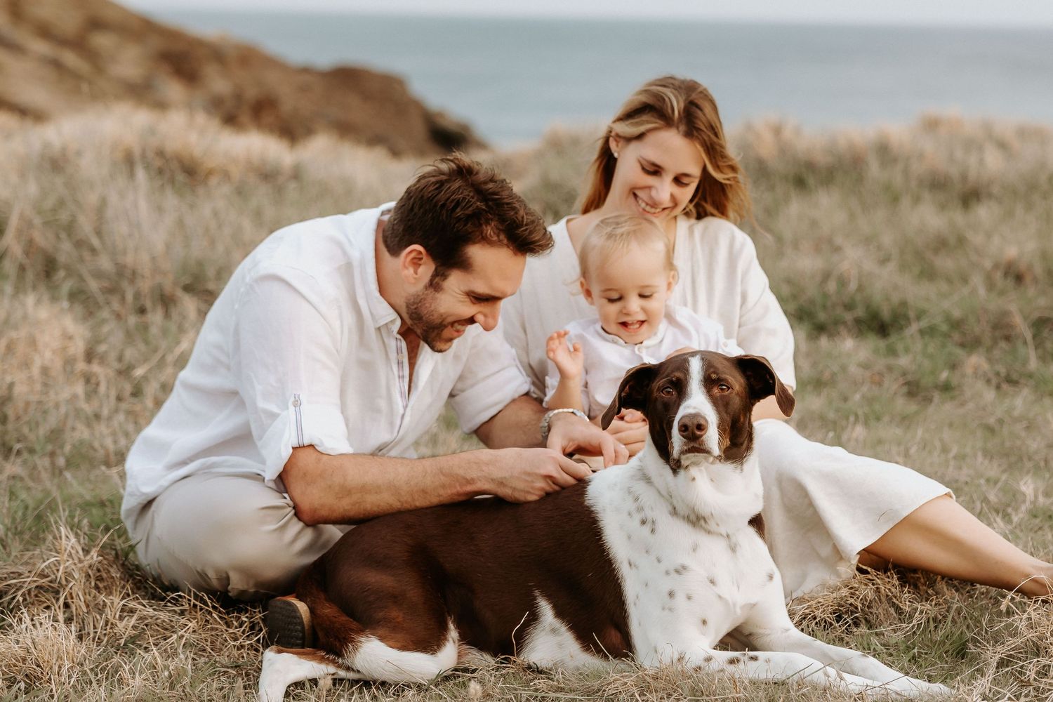 Family in white clothing sitting on grass with their dog enjoying a warm outdoor photo session together.
