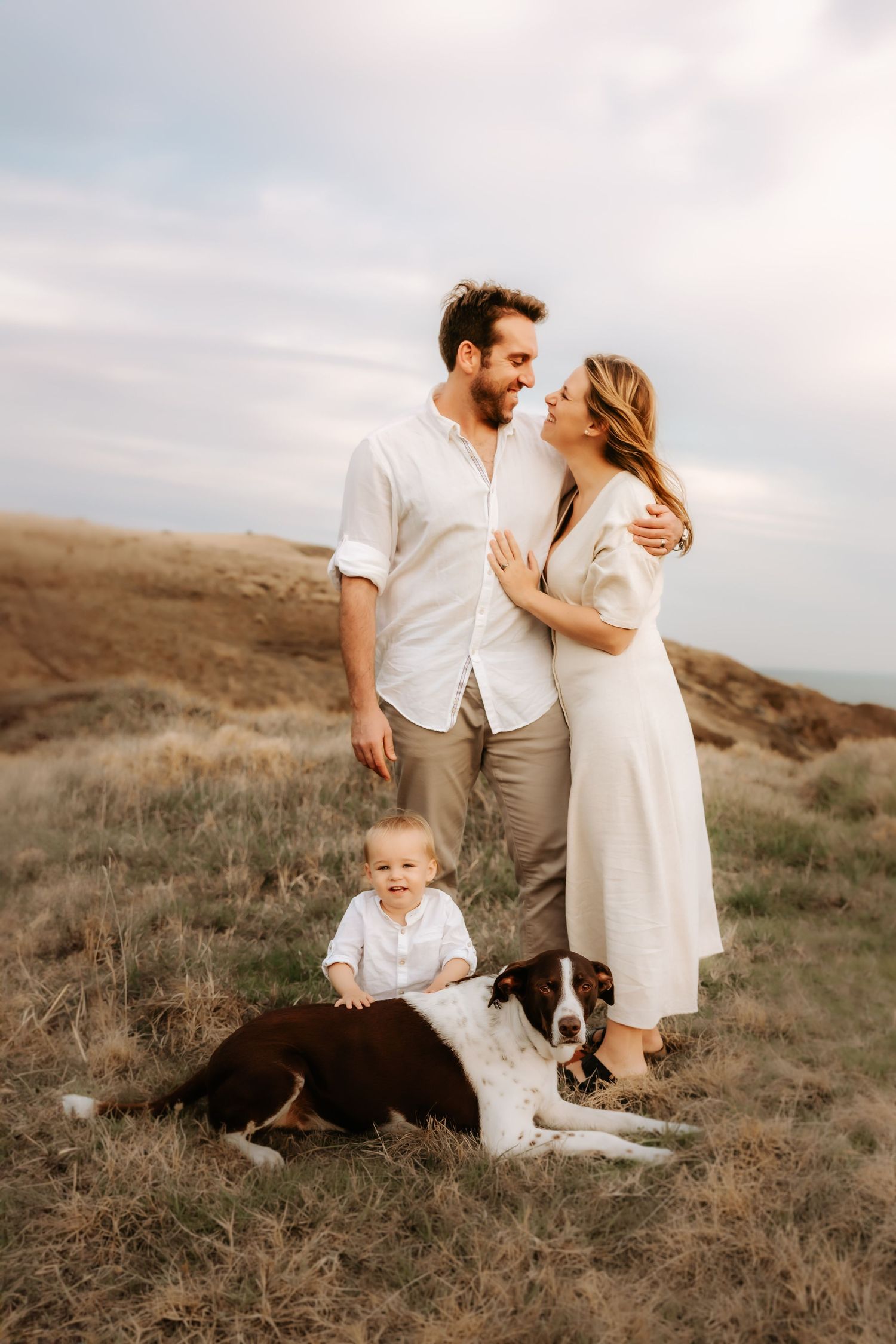 Family with young child and black and white dog posing together on hillside during sunset photoshoot.