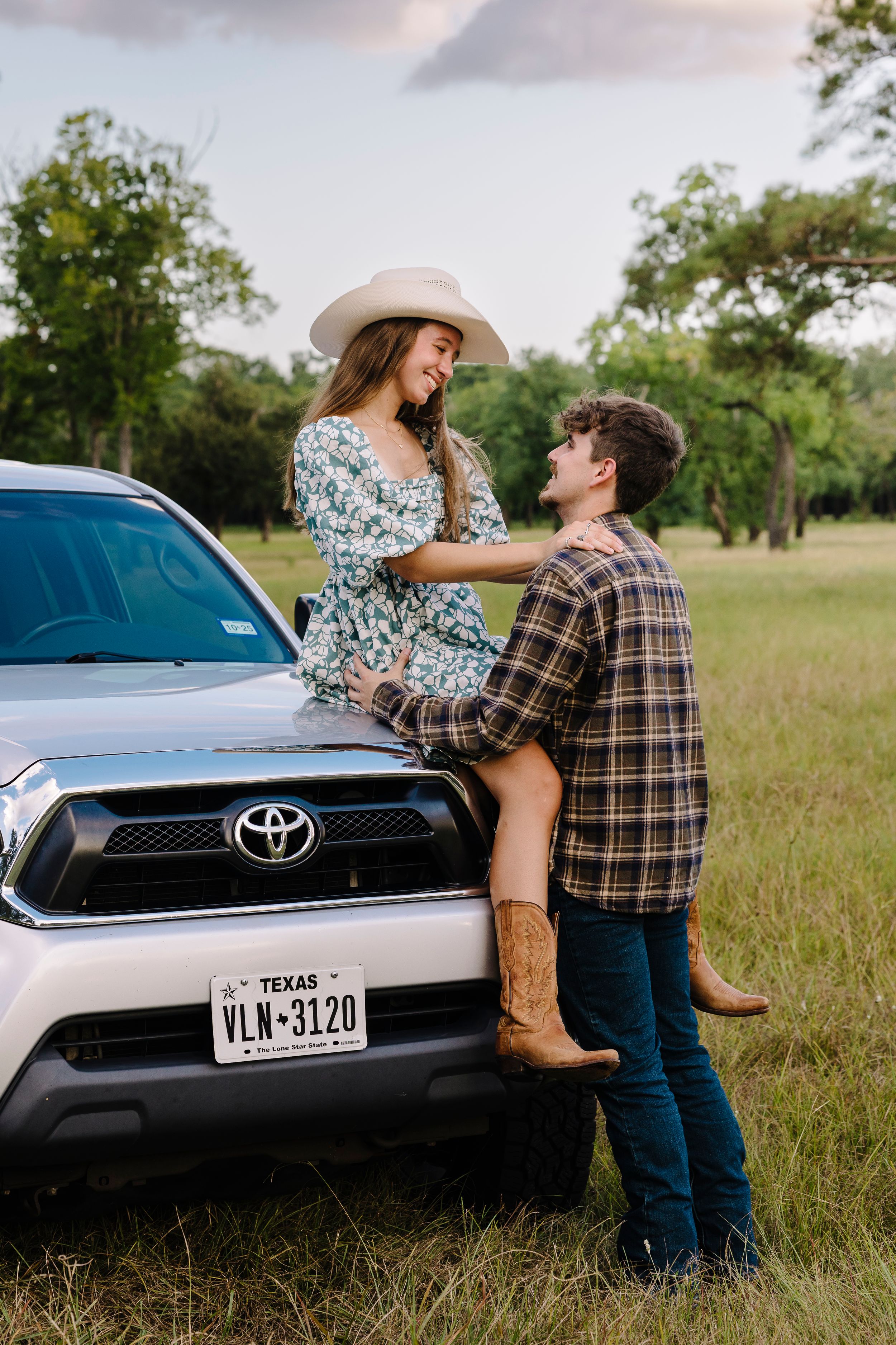 A couple share an intimate moment leaning against a blue Toyota car in a serene Texas field during sunset.