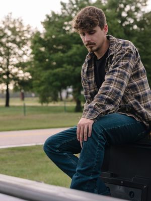 Person in plaid flannel shirt and jeans sits thoughtfully on outdoor ledge in park setting with trees at sunset.