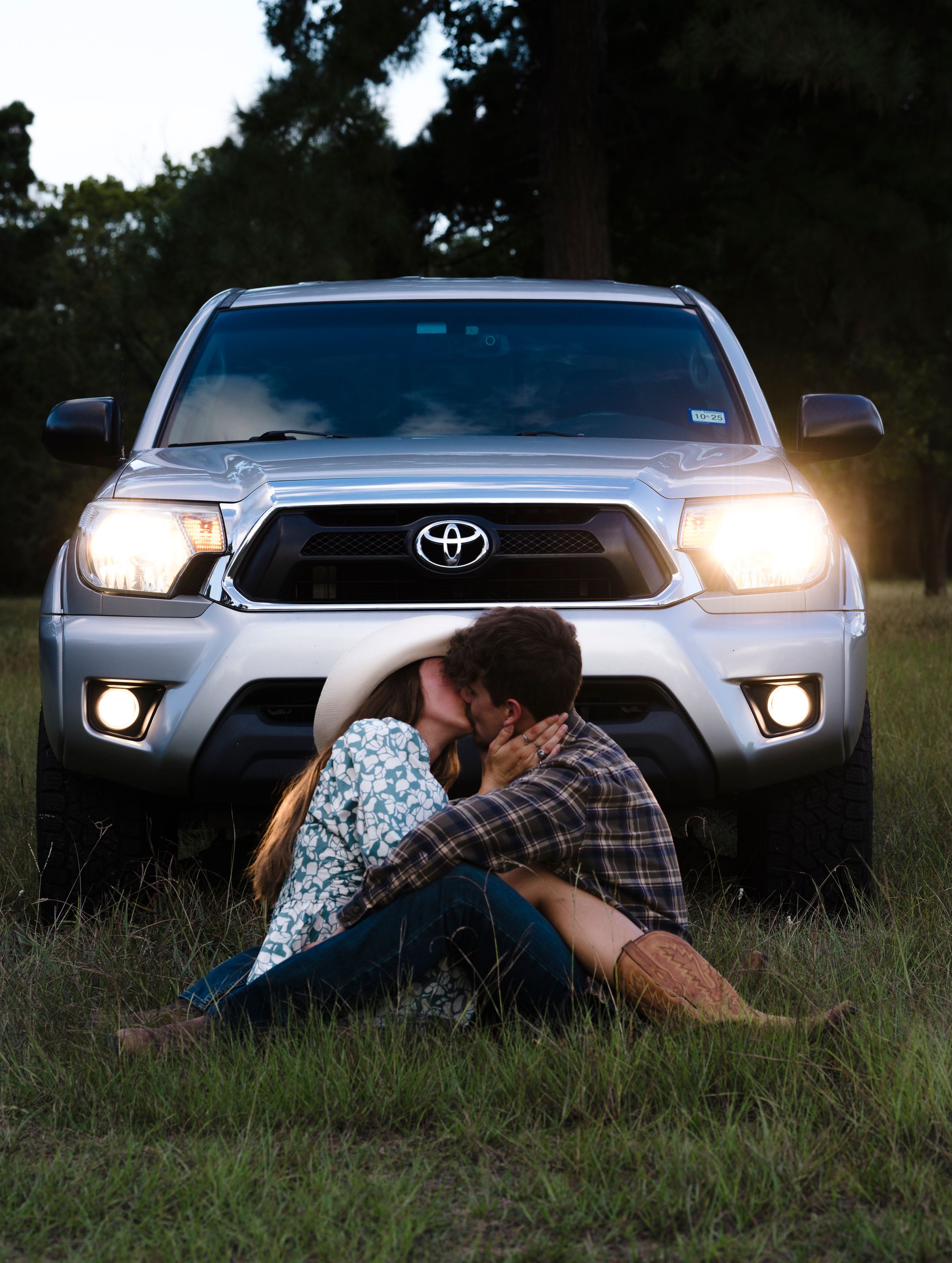 A silver Toyota Tacoma truck with illuminated headlights parked on grass during dusk while a couple sits in front.