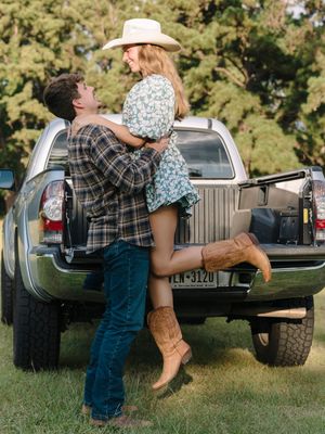 A romantic couple playfully interacts near a vintage black pickup truck on a sunny afternoon in a grassy field.