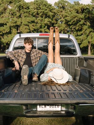 Casual couple relaxing in pickup truck bed with cowboy hat and rustic scenery.