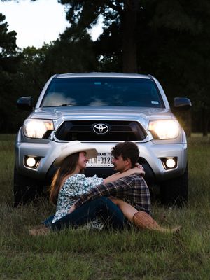A couple poses romantically in front of a Toyota pickup truck with headlights on at dusk in a grassy field.