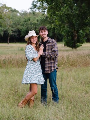 Couple in country western attire poses together in a field with trees in the background.
