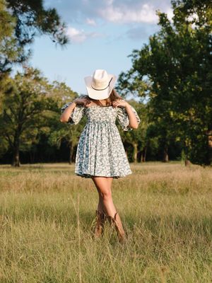 A figure in a white dress and cowboy hat walks through a sunny meadow on a summer day.