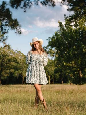 Woman in floral dress and cowboy hat poses in sunny meadow with trees.