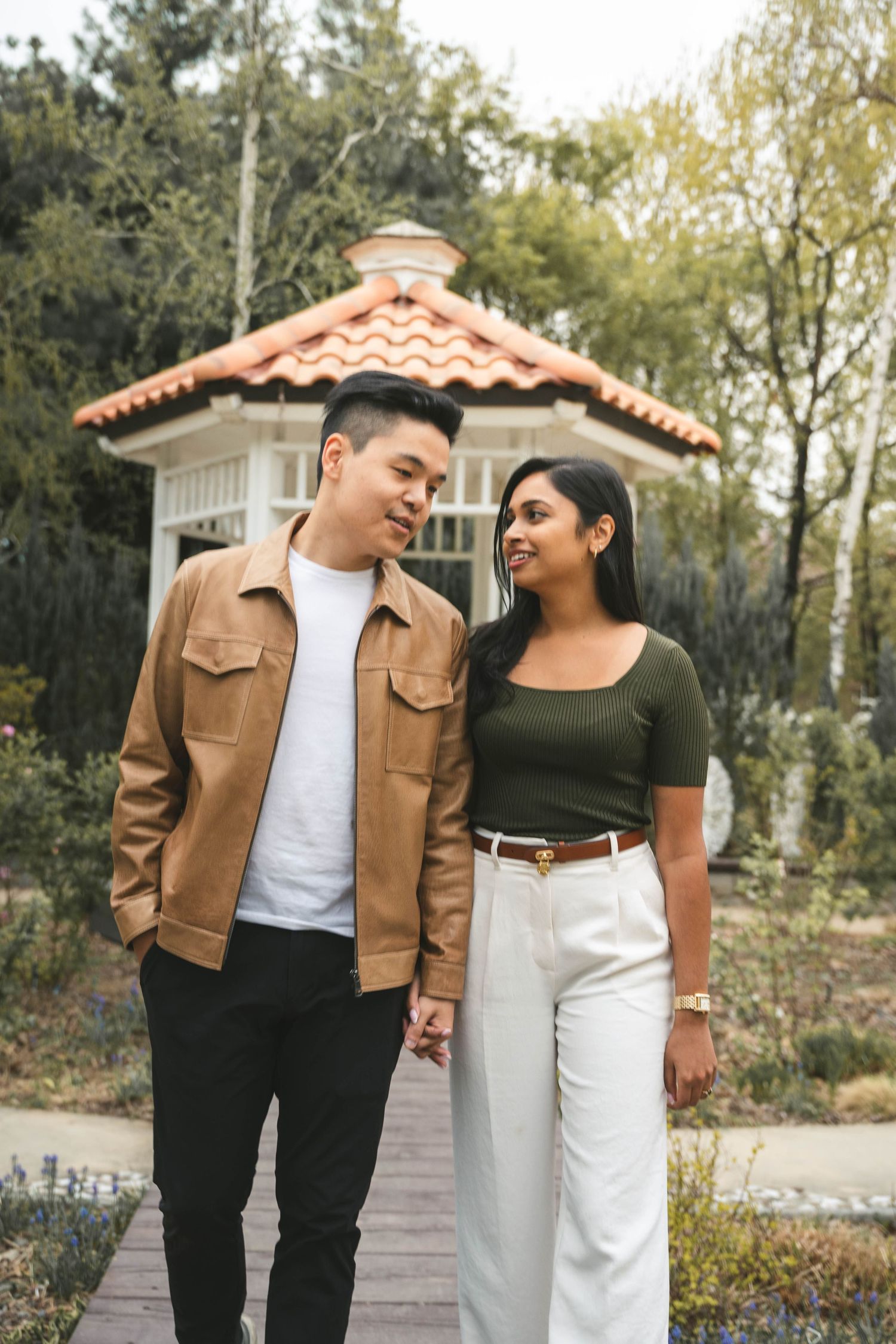 A couple in casual fall outfits walking together on a garden path with Spanish-style architecture in the background.