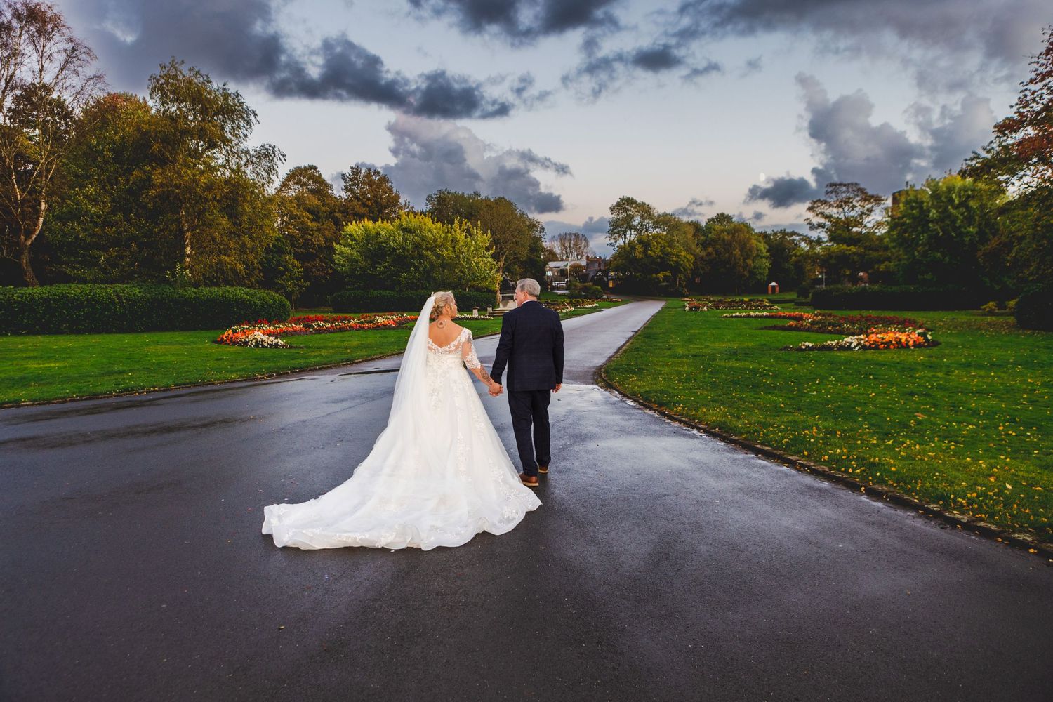 Couple walking down a winding road together through autumn trees and green lawns in cloudy weather.